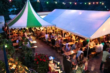 Ein lebhaftes Festzelt mit vielen Gästen, die an langen Tischen sitzen. Die Atmosphäre ist festlich, beleuchtet durch bunte Lichter.