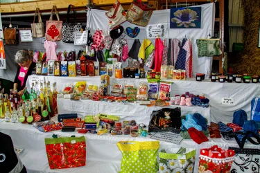 Ein Markstand voller bunter Produkte, darunter Lebensmittel und handgefertigte Taschen. Im Hintergrund ist eine Person zu sehen, die mit den Artikeln beschäftigt ist.