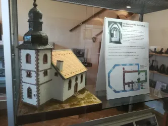 Ein Modell einer Kirche mit einem Turm und einem Satteldach steht in einer Vitrine. Daneben befindet sich ein Informationsblatt mit einem Grundriss der Kirche.