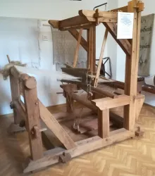 Ein traditioneller Webstuhl aus Holz steht in einem Raum. Im Hintergrund sind Stoffe und Werkzeuge sichtbar.