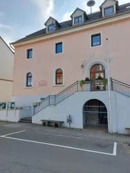 Ein modernes Rathaus mit einer hellrosa Fassade und mehreren Fenstern. Die Eingangstreppe ist von Pflanzen umgeben und es gibt eine Straßensignalisierung vor dem Gebäude.