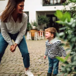Eine lächelnde Frau spielt fröhlich mit einem kleinen Jungen im Freien. Im Hintergrund sind grüne Pflanzen und eine gemütliche Atmosphäre.
