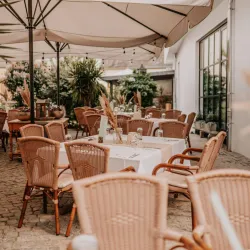 Ein stilvolles Café im Freien mit wicker Stühlen und eleganten Tischen. Der Raum ist von Pflanzen umgeben und hat ein einladendes Ambiente.