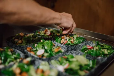 Frische Salate werden in Schalen angerichtet. Eine Hand fügt zuletzt Zutaten hinzu.