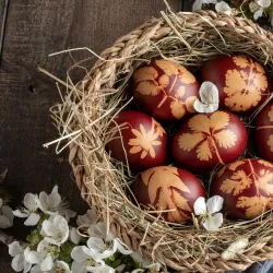 Ein Korb mit dekorierten Ostereiern in verschiedenen rötlichen Tönen liegt auf einem Holztisch. Um den Korb herum befinden sich frische Blumen und Gras.