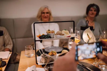 Ein stilvoll angerichteter Tisch mit einem mehrstöckigen Servierstand voller köstlicher Speisen. Im Hintergrund sind zwei Personen zu sehen, die entspannt am Tisch sitzen.