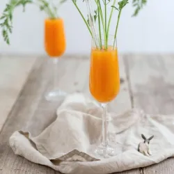 Zwei Gläser mit orangefarbigem Getränk, dekoriert mit Karottenblättern. Sie stehen auf einem hellen Tisch, umgeben von einem neutralen Tuch.