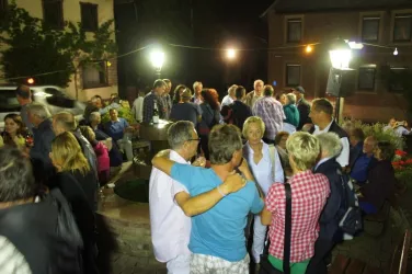 Eine fröhliche Menschenmenge bei Nacht versammelt sich um einen Brunnen. Die Atmosphäre ist lebhaft, mit Musik und Lichtern.