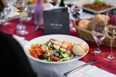 Ein Teller mit frischem Salat, Garnelen und einer Brötchenhälfte. Im Hintergrund sind Gläser mit Wein und eine festlich gedeckte Tafel zu sehen.