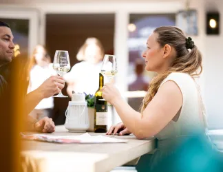 Ein Paar hebt Gläser mit Wein an einem Tisch an. Im Hintergrund sind weitere Personen zu sehen, die eine entspannte Atmosphäre genießen.