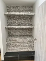 Ein Regal mit Backsteinmuster an der Wand und zwei offenen Regalen. Der Boden ist mit dunklen Fliesen gestaltet.