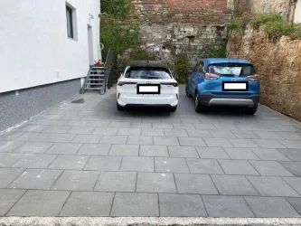 Ein Parkplatz mit zwei Autos, einem weißen und einem blauen. Im Hintergrund ist eine Wand aus Stein und eine Treppe zu sehen.