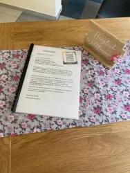 Ein Holzschild mit dem Text "Herzlich Willkommen" steht neben einem bedruckten Blatt in einem Klarsichtordner. Der Tisch ist mit einer blumigen Tischdecke dekoriert.