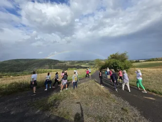Eine Gruppe von Menschen wandert auf einem Weg in der Natur. Im Hintergrund ist ein Regenbogen über sanften Hügeln und einem grünen Feld sichtbar.