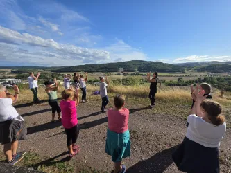 Eine Gruppe von Menschen macht Übungen im Freien mit Blick auf eine malerische Landschaft. Der Himmel ist blau und es sind einige Wolken zu sehen.