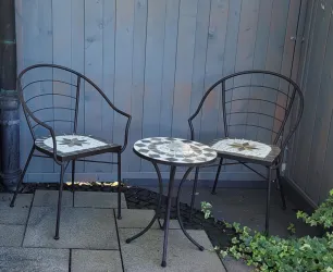 Zwei schmiedeeiserne Stühle mit gemusterten Sitzflächen stehen um einen kleinen runden Tisch. Der Hintergrund ist eine hölzerne Wand und es gibt Pflanzen am Boden.