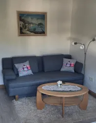 Ein gemütliches Wohnzimmer mit einem grauen Sofa und Kissen mit Eulenmotiven. Ein runder Holztisch steht vor dem Sofa, und an der Wand hängt ein Landschaftsgemälde.