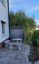 Ein kleiner Gartenbereich mit einem Steingartentisch und zwei Stühlen. Im Hintergrund stehen grüne Pflanzen und ein Sichtschutz.