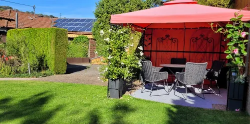 Ein schöner Garten mit einem roten Pavillon und bequemen Stühlen. Im Hintergrund sind Grünanlagen und ein blauer Himmel zu sehen.
