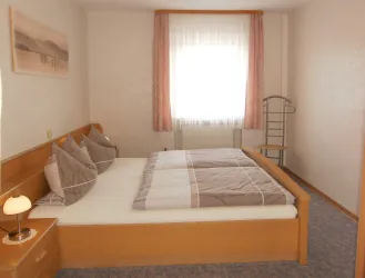 Ein gemütliches Schlafzimmer mit einem Doppelbett und zwei Kissen. Das Zimmer hat helles Licht von einem großen Fenster und eine ruhige Atmosphäre.