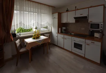 Eine helle Küche mit einem Tisch und Stühlen. Im Hintergrund steht ein Fenster mit Pflanzen und Küchengeräten.