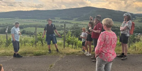 Eine Gruppe von Menschen steht im Freien, während sie einem Mann zuhören, der ihnen etwas erklärt. Im Hintergrund sind sanfte Hügel und Weinreben zu sehen.