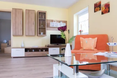 Ein modernes Wohnzimmer mit einer orangen Couch und einem Glaskaffee-Tisch. An den Wänden hängen einige Bilder und es gibt Pflanzen sowie einen Fernseher.