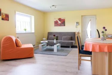 Ein gemütliches Wohnzimmer mit einem orangen Sessel und einem grauen Sofa. Der Raum hat gelbe Wände und einen Esstisch mit Stühlen.