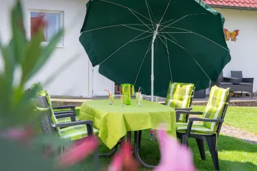 Ein gepflegter Garten mit einem grünen Sonnenschirm und einem Tisch, der mit grüner Tischdecke und Stühlen gedeckt ist. Auf dem Tisch stehen zwei Gläser, die einladend wirken.