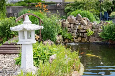 Ein schöner Garten mit einem Teich und einem Steinwasserfall. Eine japanische Laterne steht neben üppigem Grün und farbigen Fischen im Wasser.