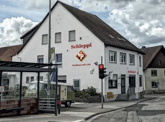 Ein weißes Gebäude mit dem Schriftzug "Schleppi" auf der Fassade. In der Nähe steht eine Haltestelle und ein Verkehrsampel zeigt Rot.