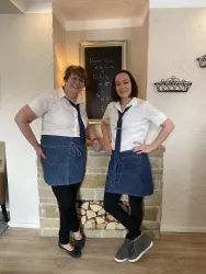 Zwei Damen stehen lächelnd vor einem Kamin. Sie tragen einheitliche Outfits mit weißen Shirts und blauen Schürzen.