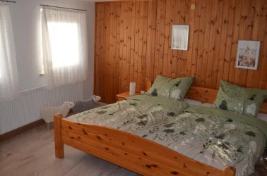 Ein gemütliches Schlafzimmer mit einem Holzbett und einer grünen Bettwäsche. An den Wänden sind helle Fenster und ein Holzpanel zu sehen.