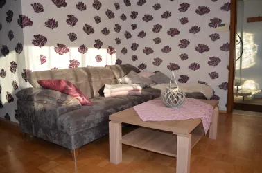 Ein gemütliches Wohnzimmer mit einem grauen Sofa und einem Holztisch. Die Wände sind mit einem floralen Muster dekoriert.