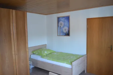 Ein schlichtes Schlafzimmer mit einem Einzelbett und einer grünen Bettdecke. An der Wand hängt ein Bild von Löwenzahnblüten.