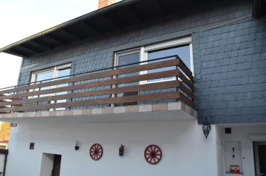 Ein modernes Haus mit einem Holz-Balkon und großen Fenstern. Die Fassade ist teilweise aus grauen Schieferplatten und weißen Wänden mit dekorativen Elementen.
