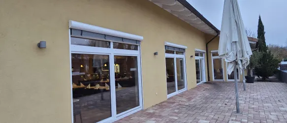 Ein modernes Gebäude mit großen Fenstern und einer gelben Fassade. Auf dem Bürgersteig stehen Sonnenschirme und Pflanzen.