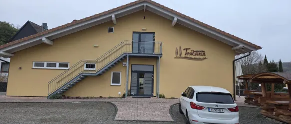 Ein modernes, gelbes Gebäude mit einer geräumigen Treppe und großen Fenstern. Vor dem Eingang steht ein weißes Auto auf einem gepflasterten Platz.