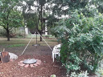 Ein Garten mit mehreren Bäumen und einem Feuerplatz. Im Vordergrund steht eine Schwinge und ein weißer Stuhl.
