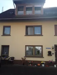 Ein zweistöckiges Haus mit gelber Fassade und Fenstern. Vor dem Haus stehen Blumenkübel und zwei Stühle.