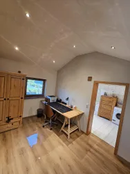 Ein helles Arbeitszimmer mit einem Schreibtisch, einem Stuhl und einem Schrank aus Holz. Durch das Fenster ist eine grüne Landschaft zu sehen.