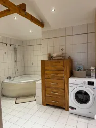 Ein modernes Badezimmer mit einer Badewanne und einer Waschmaschine. Es gibt einen Holzschrank und helle Fliesen.