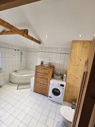 Ein modernes Badezimmer mit einer Badewanne, einer Waschmaschine und einem Holzschrank. Helle Fliesen und eine offene Raumgestaltung sorgen für eine freundliche Atmosphäre.