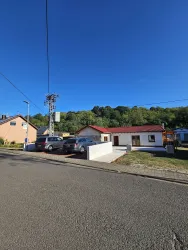 Ein ruhiges Wohnviertel mit einem einfachen Haus und einem klaren blauen Himmel. Im Vordergrund stehen mehrere Autos am Straßenrand.