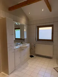 Ein helles Badezimmer mit weißen Fliesen und einem modernen Waschbecken. Große Fenster sorgen für viel Tageslicht.