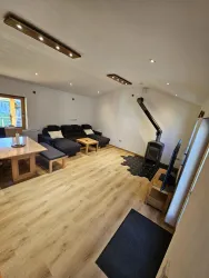 Ein helles Wohnzimmer mit einer gemütlichen Couch und einem Holzofen. Der Boden ist aus hellem Holz und es gibt einen großen Esstisch.