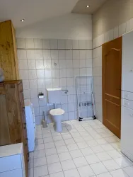 Ein helles Badezimmer mit weißen Fliesen und einem WC. Im Raum gibt es einen Holzschrank und einen Wäscheständer.