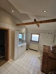 Ein modernes Badezimmer mit weißen Fliesen und Holzdetails. Es gibt ein großes Fenster und eine typischen Ausstattung mit Waschtisch und Spiegel.