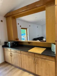 Eine moderne Küche mit Holzmöbeln und einem schwarzen Arbeitsbereich. Im Hintergrund sieht man einen Wohnbereich mit einem Sofa.