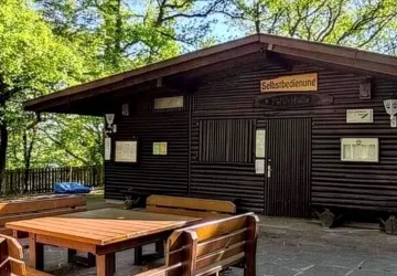 Eine gemütliche Selbstbedienungshütte umgeben von Bäumen. Im Vordergrund stehen Holztische und -bänke.